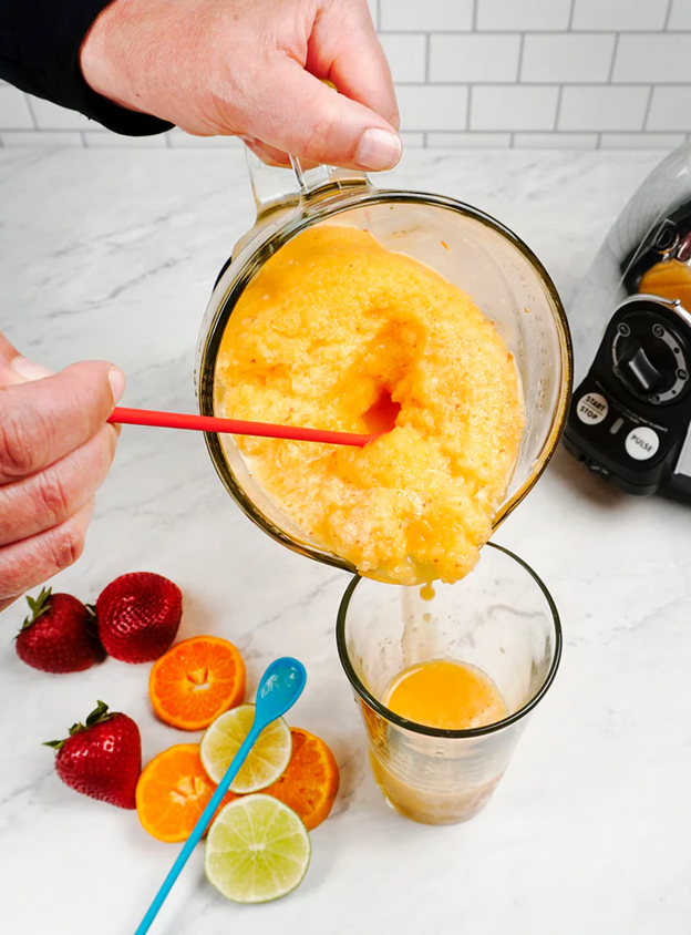 long handle silicone spoon scooping smoothie out of blender into glass with strawberries, lemon slices and orange slices on kitchen counter