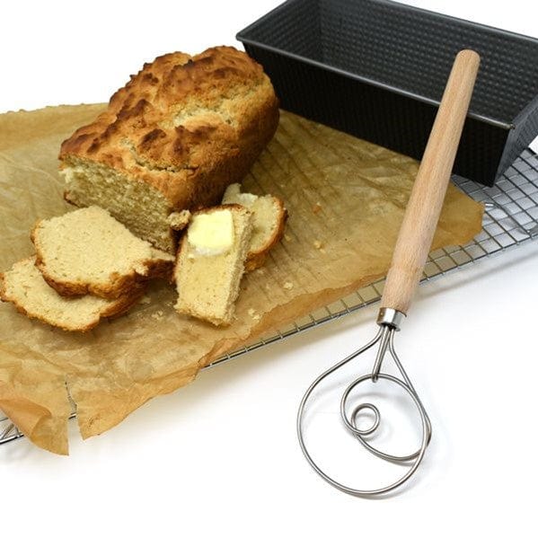 Norpro 12'' Danish Dough Whisk in front of bread pan and partially cut loaf of sourdough bread with slices of sourdough bread sitting on cooling rack and a slab of butter on top