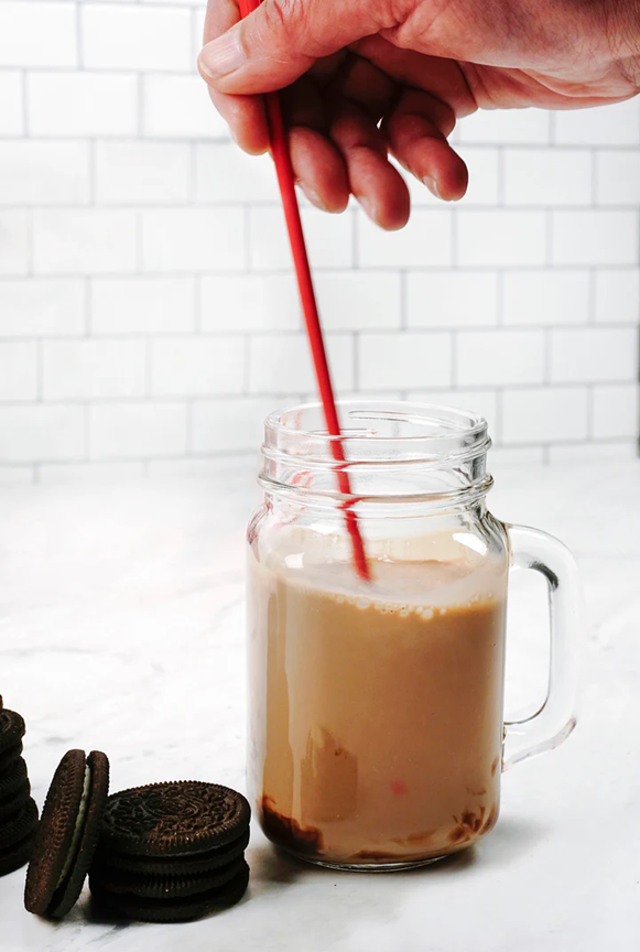 red silicone stirring spoon mixing a milkshake in a glass cup with handle on kitchen counter next to chocolate cookies